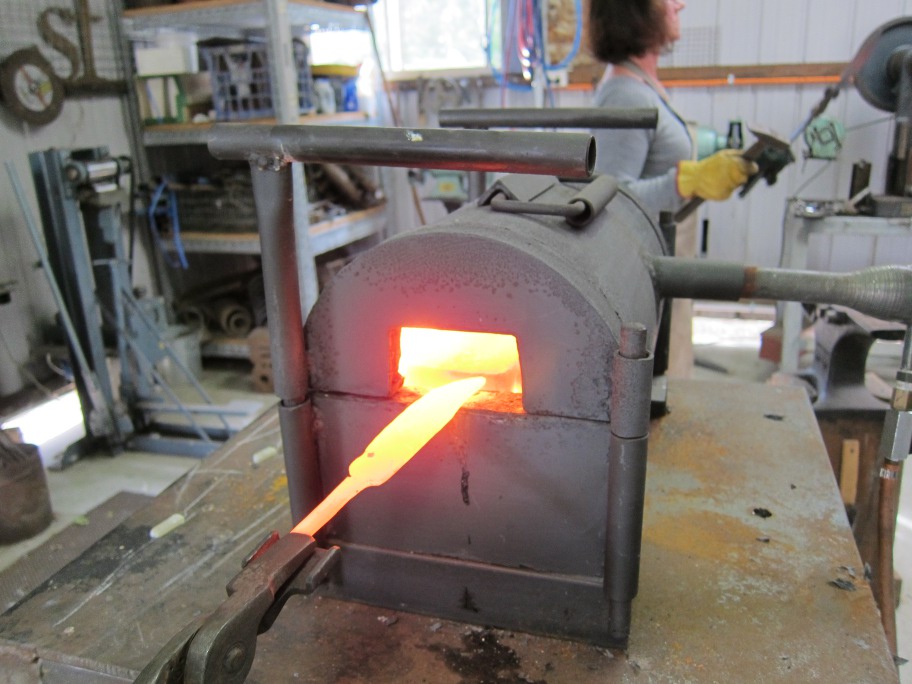 A  bright yellow bar of metal with a knife shape on one end is withdraw from the gas forge.